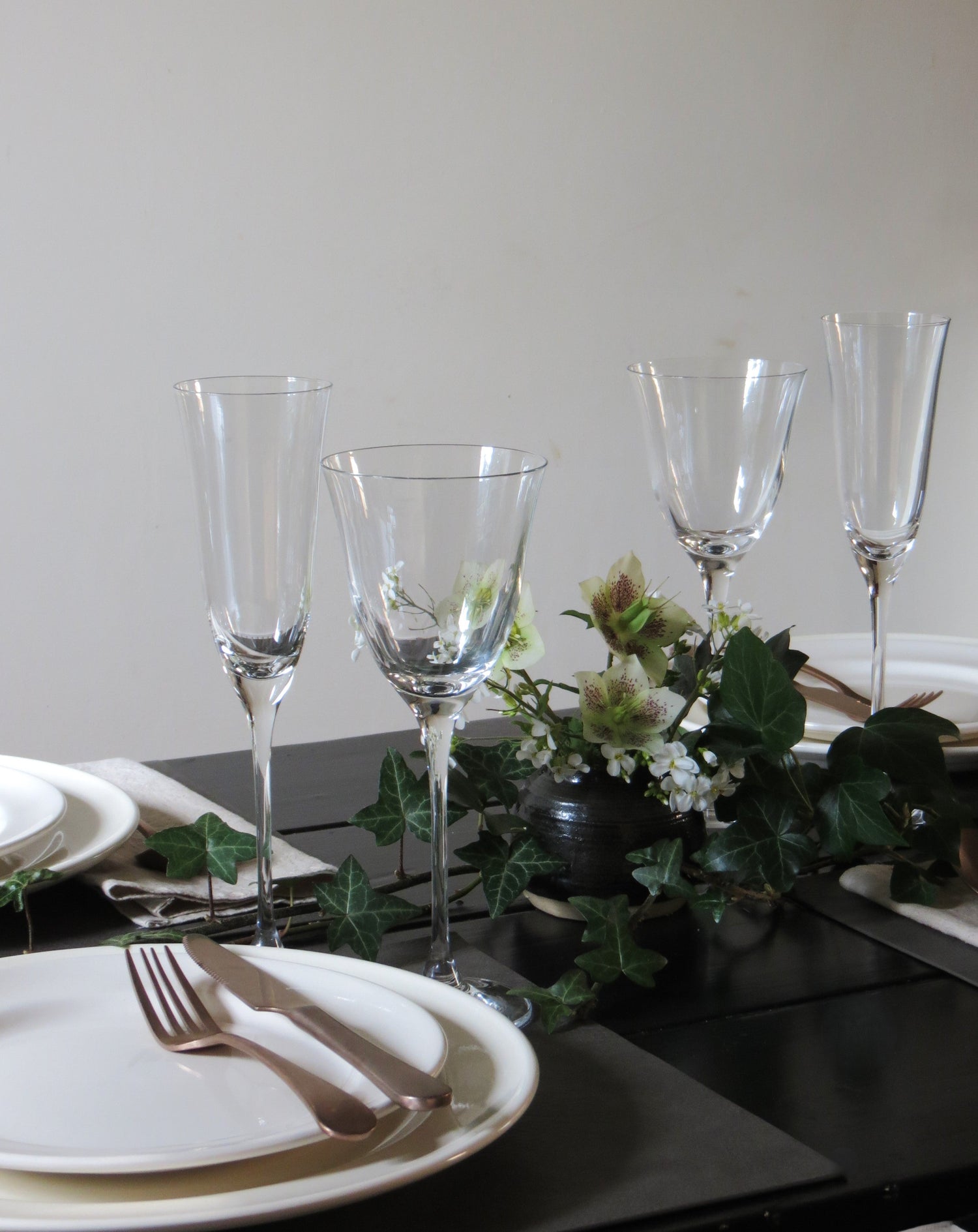 Dark table with ivory plates and a floral arrangement in the middle. Tulip shaped glasses stand out on the setting.  
