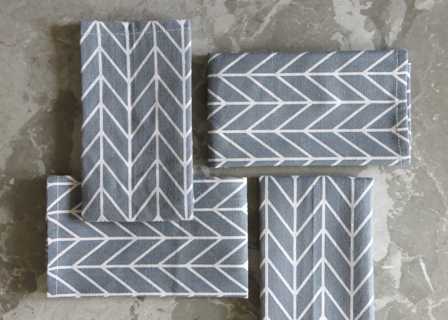 A set of four folded grey napkins with a white chevron pattern, displayed on a grey surface.
