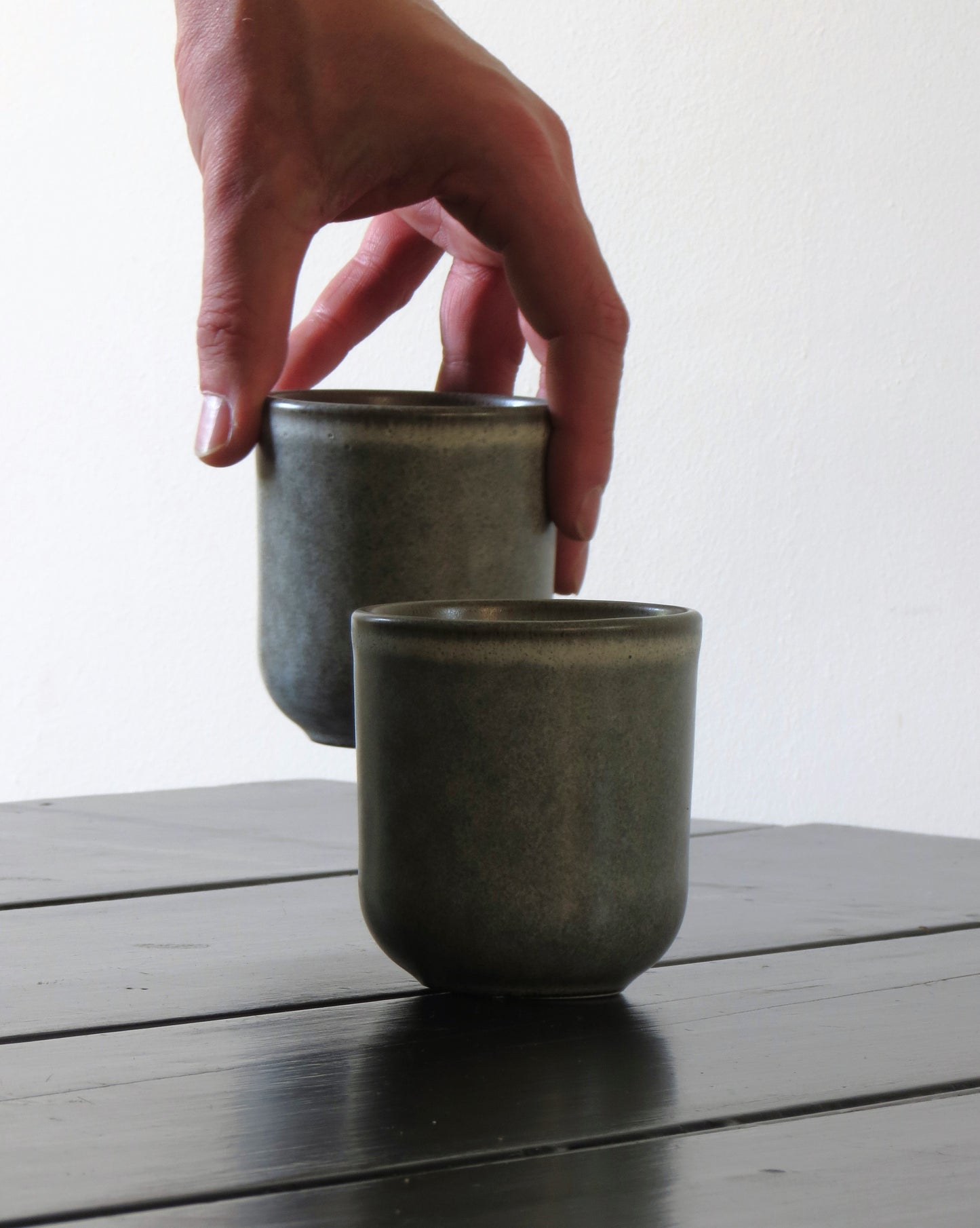 Two dark ceramic cups on a wooden surface with a hand reaching towards one of them.