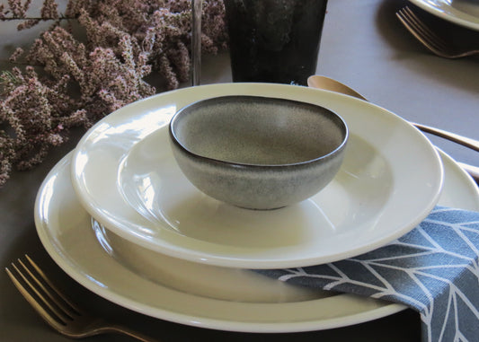 A beige Clay Mini Bowl with a matte finish, placed inside a larger white plate, with a blue and white patterned napkin to the side and a floral arrangement in the background.