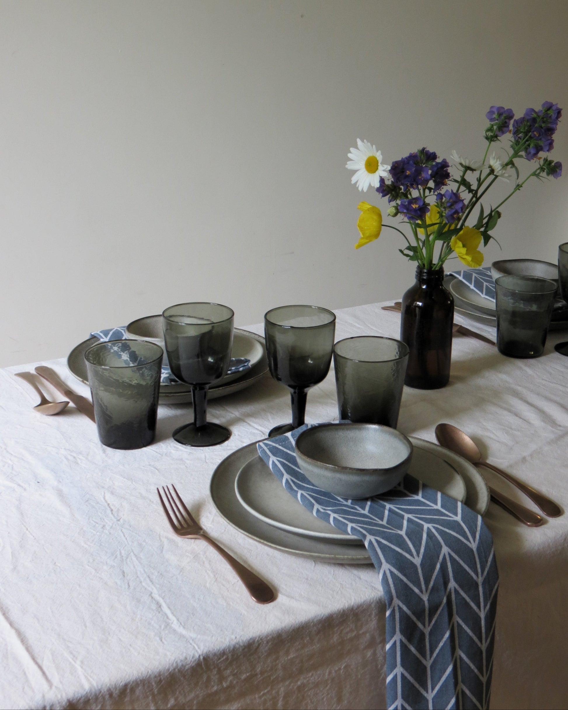Dining table set with grey glassware, plates, and cutlery on a white tablecloth.