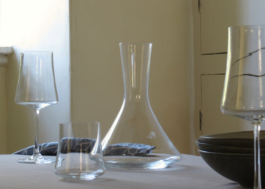 A clear glass decanter with a sleek design, displayed on a table with two empty glasses and a bowl in the background.