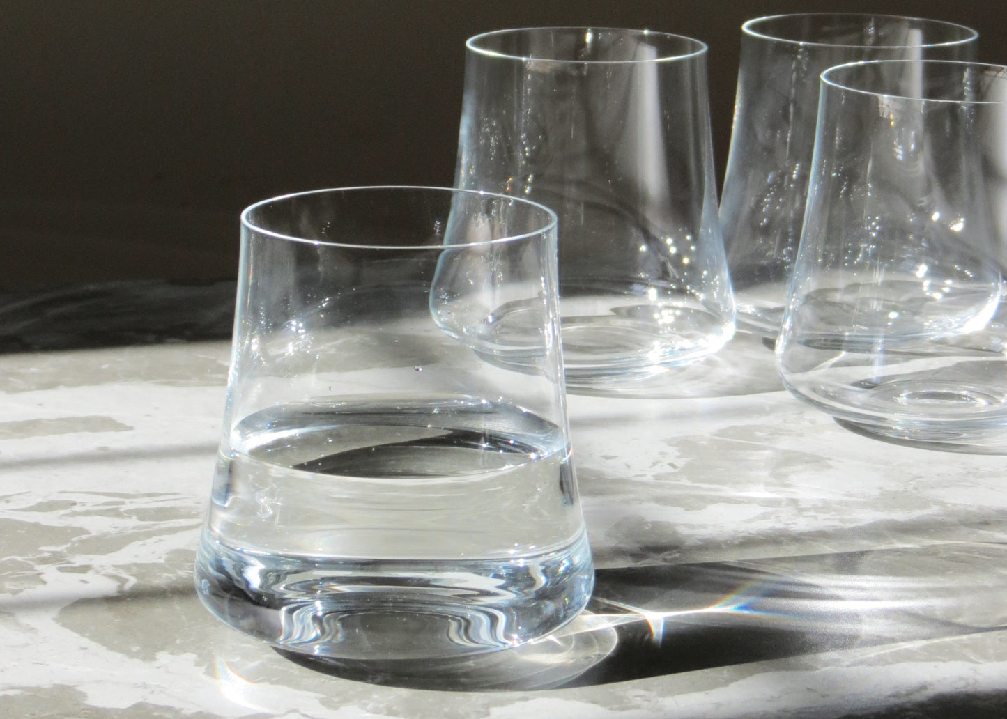 A set of four clear crystalline glass tumblers on a light surface, with a full glass in the foreground and three empty tumblers in the background.