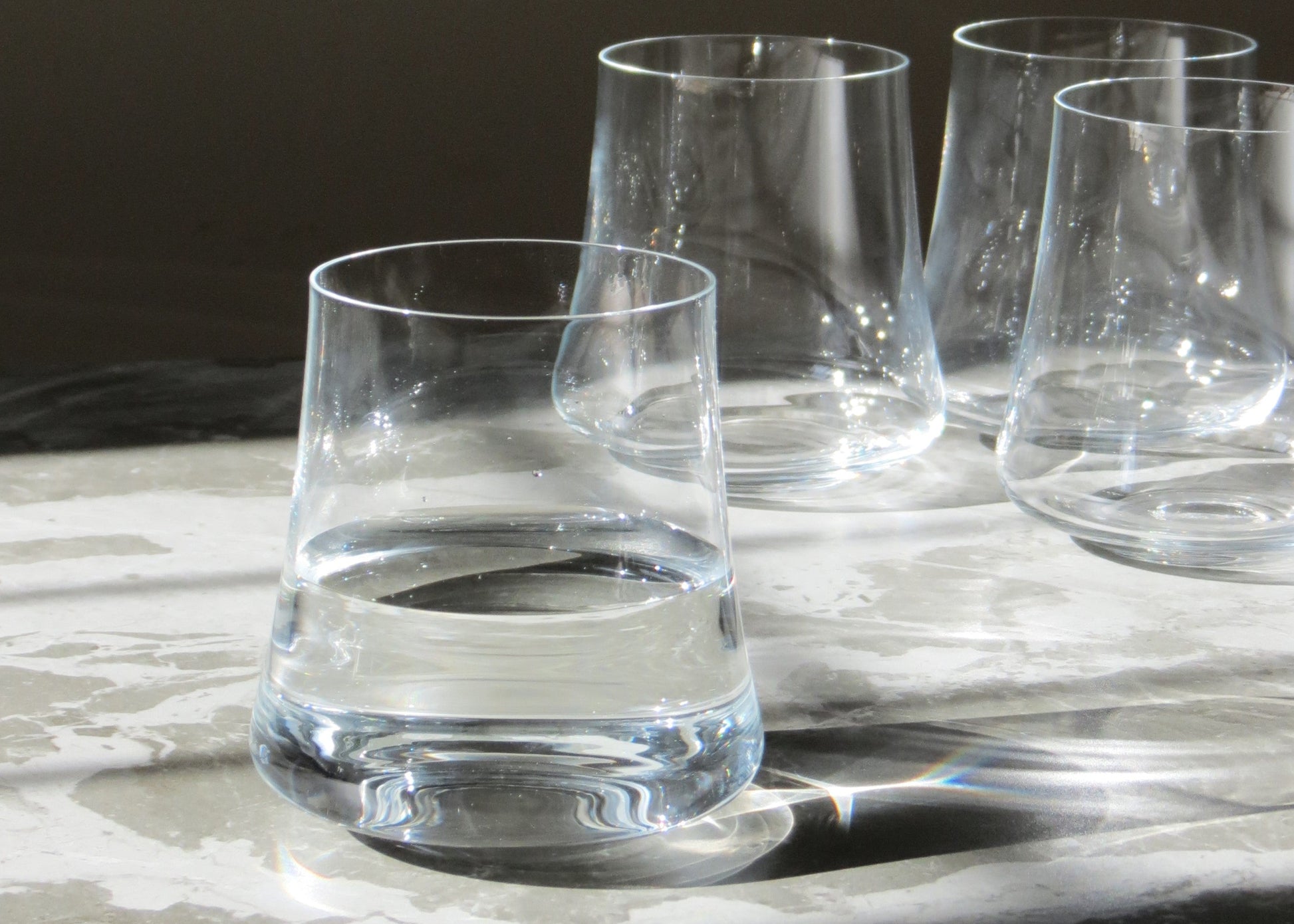 A set of four clear crystalline glass tumblers on a light surface, with a full glass in the foreground and three empty tumblers in the background.