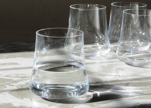 A set of four clear crystalline glass tumblers on a light surface, with a full glass in the foreground and three empty tumblers in the background.