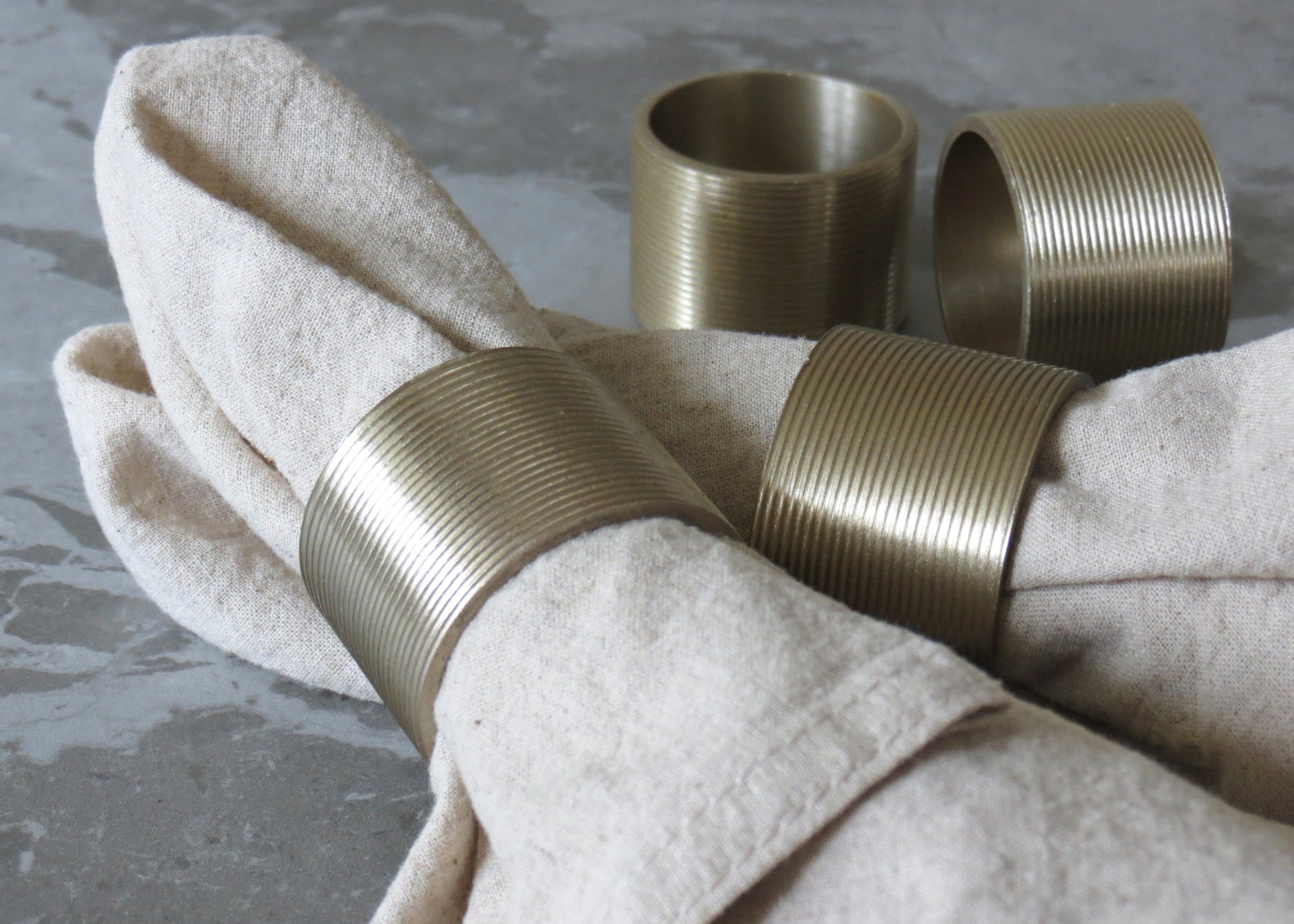 A set of four gold-colored lined napkin rings displayed on a beige napkin, with a grey background.