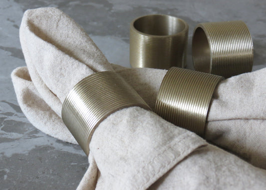 A set of four gold-colored lined napkin rings displayed on a beige napkin, with a grey background.
