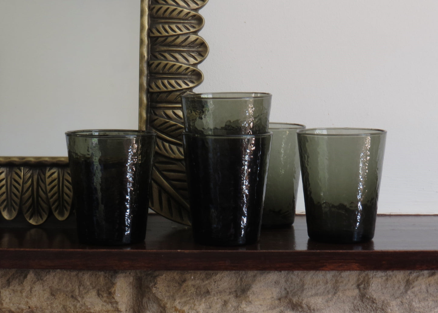Four smoked glass tumblers on a wooden surface with a decorative mirror in the background.