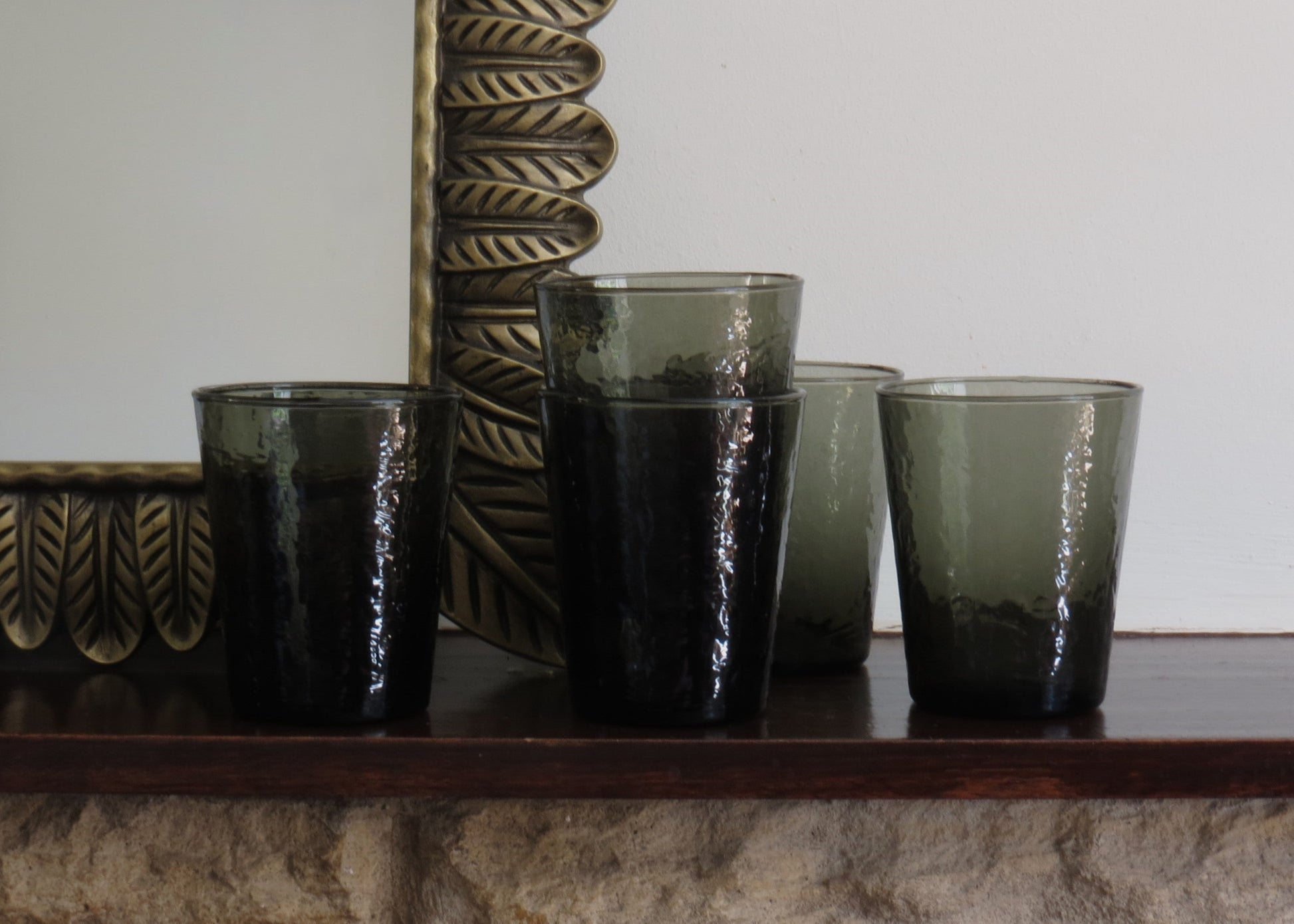 Four smoked glass tumblers on a wooden surface with a decorative mirror in the background.