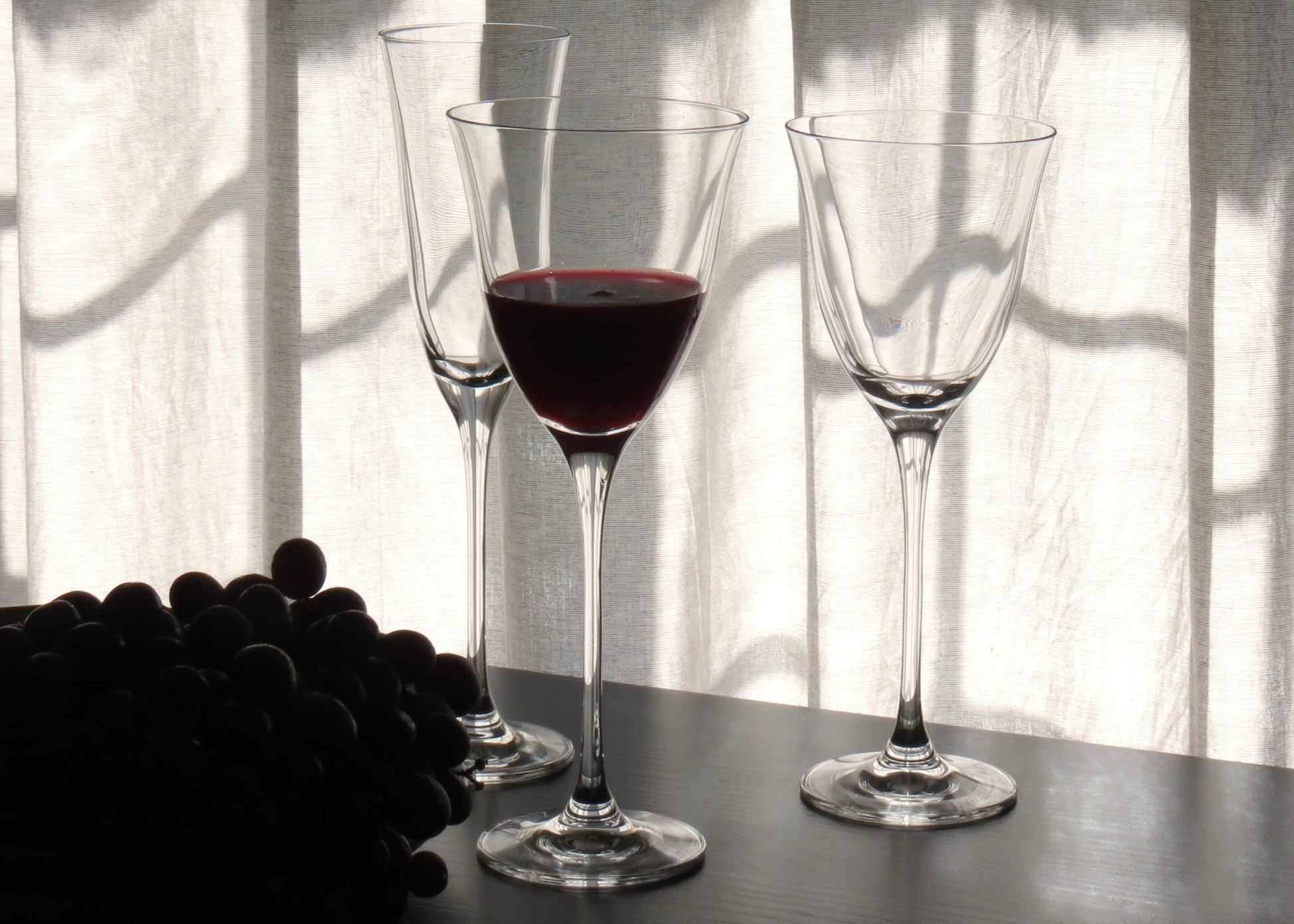 Two wine glasses with red wine on a table next to grapes, with sunlight filtering through a curtain.